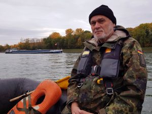 Der Stabsunteroffizier Heinz Cecil befindet sich mit einem Sturmboot der Bundeswehr auf dem Rhein. Im Hintergrund ist ein Binnenschiff zu sehen.